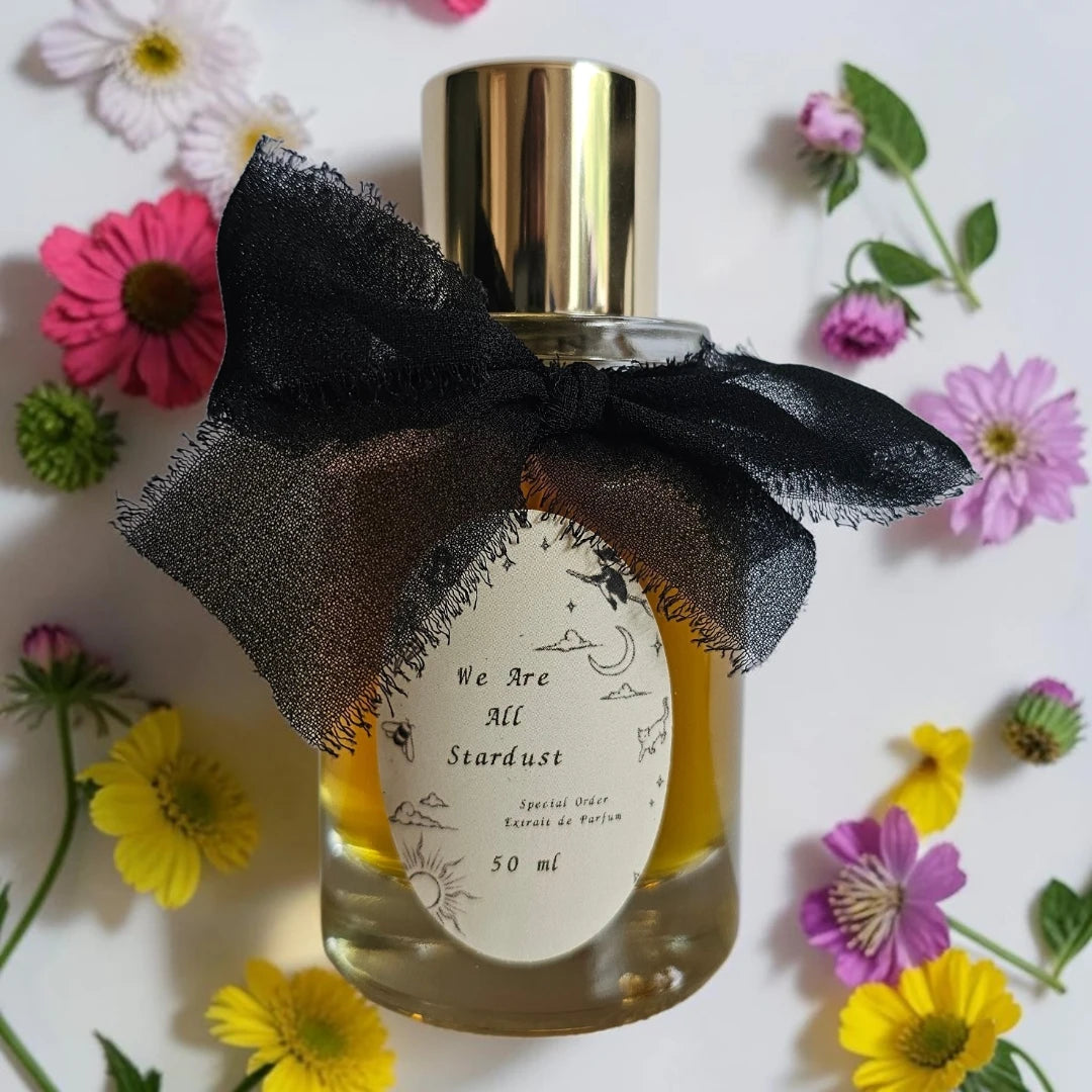 A 50 ml bottle of 'We Are All Stardust' special edition scented oil, decorated with a black sheer ribbon bow and a paper label, surrounded by colorful artificial flowers on a white background.
Sorce we are all stardust | Fragrance Samples UK available in 1ml 2ml 5ml samples 
