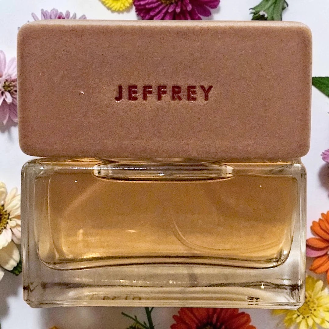 Jeffrey Garrett perfume bottle with a tan rectangular cap embossed with the brand name, filled with amber liquid, set against a white background with colorful flowers.