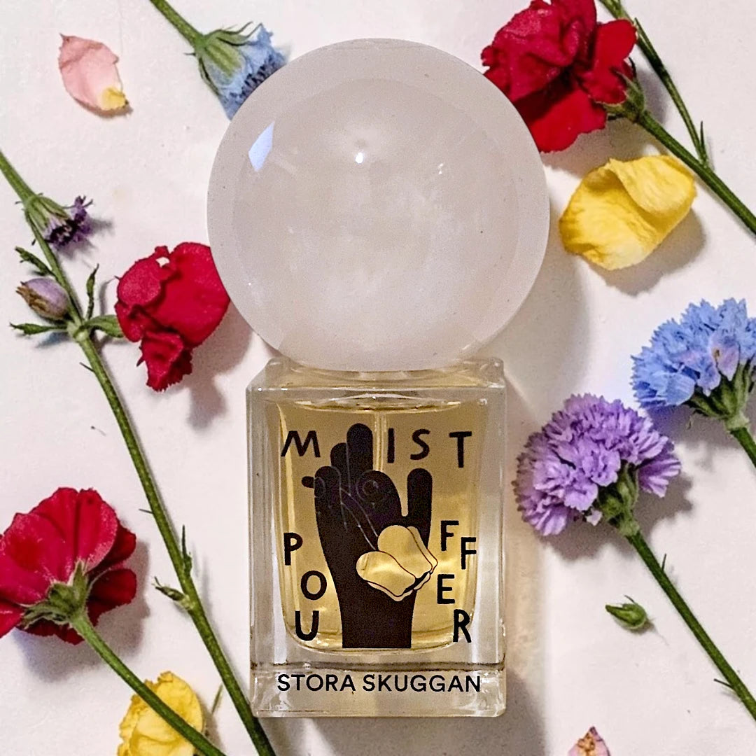 A perfume bottle labeled 'Mist Pour' by Stora Skuggan, featuring a large translucent white crystal sphere cap and a black hand graphic over a gold background. The bottle is surrounded by scattered red, yellow, blue, and purple flowers on a white surface.