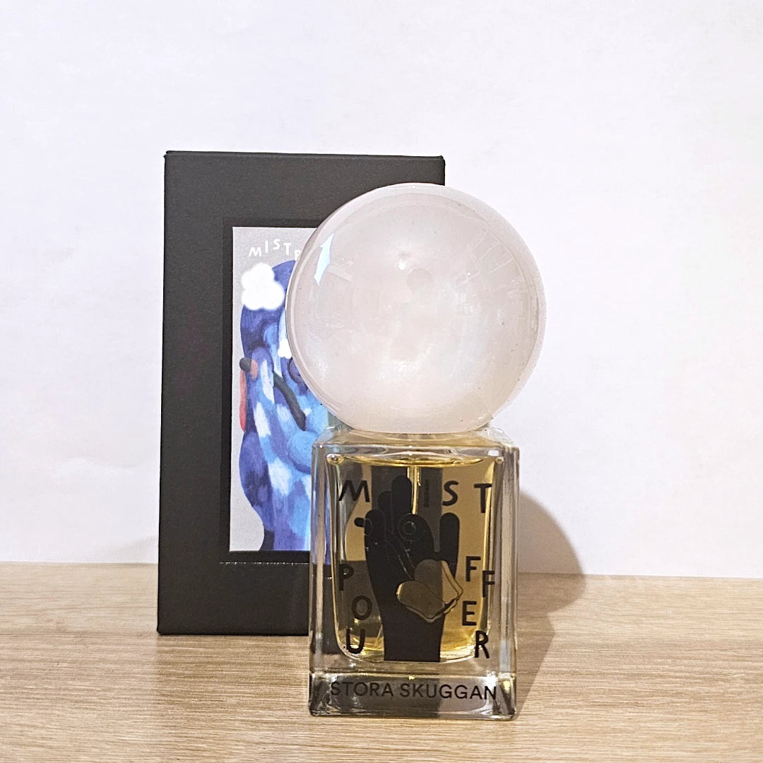 A bottle of Misty Flower perfume by Victoria Skuggan with a large white crystal sphere cap, shown in front of its black packaging box on a light wood surface against a white wall.
