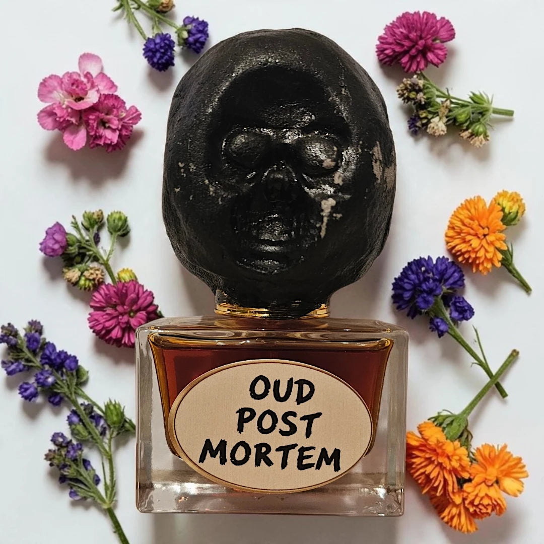 A perfume bottle with a sculpted black skull cap and a clear glass base filled with amber liquid, surrounded by small colorful flowers on a white background. The bottle label reads 'OUD POST MORTEM'.