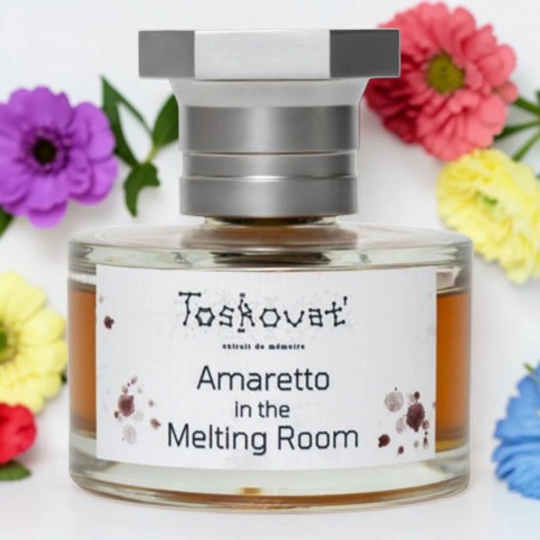 A clear glass perfume bottle with amber liquid and a silver cap, labeled 'Toskovat Amaretto in the Melting Room', surrounded by colorful flowers on a white background. Toskovat Amaretto In The Melting Room | Fragrance Samples UK