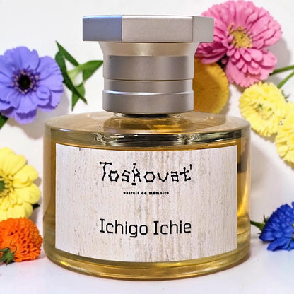 A bottle of Toskovat 'Ichigo Ichie' perfume with a silver cap and a light wood-grain label, surrounded by colorful flowers on a white background.