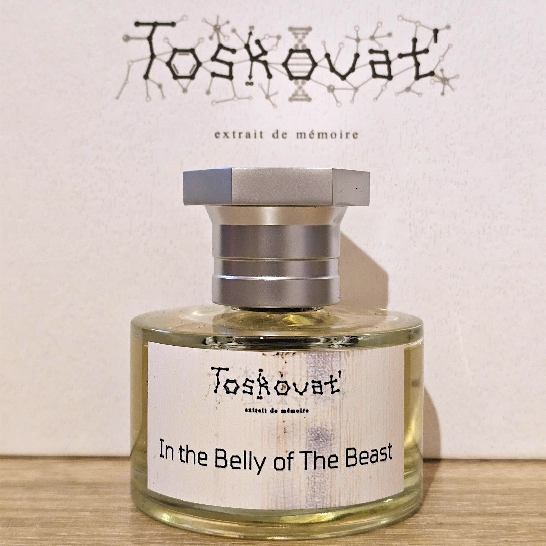 A bottle of Toskovat 'In the Belly of The Beast' extrait de mémoire perfume, featuring a clear glass bottle with pale yellow liquid, a white label, and a metallic silver cap, set against a textured off-white background with the brand name printed above.