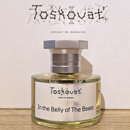 A bottle of Toskovat 'In the Belly of The Beast' extrait de mémoire perfume, featuring a clear glass bottle with pale yellow liquid, a white label, and a metallic silver cap, set against a textured off-white background with the brand name printed above.