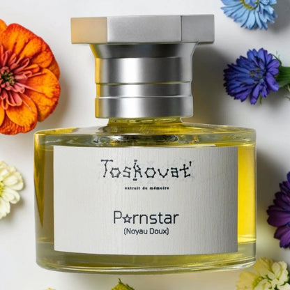 Toskovat Pornstar NOYAU DOUX fragrance sample 1ml 2ml 5ml samples available 
A bottle of Toskovat 'Pornstar' (Noyau Doux) perfume with a silver cap and a white label, filled with yellow liquid, surrounded by colorful flowers on a white background.