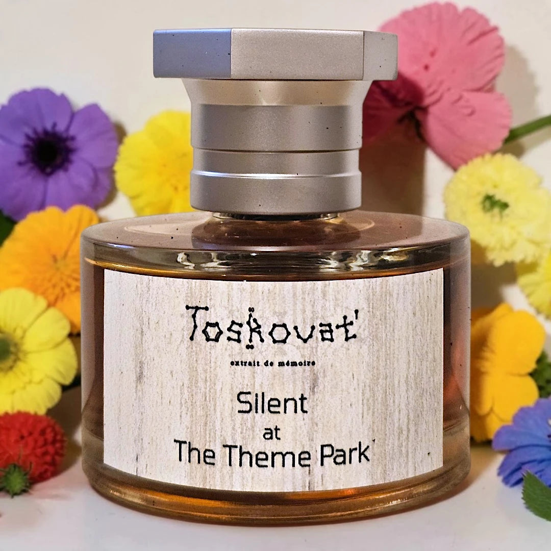 A bottle of Toskovat 'Silent at The Theme Park' memory perfume with a silver cap and a light wood-grain label, surrounded by colorful artificial flowers. Toskovat Silent at The Theme Park | Fragrance Samples UK available in 1ml, 2ml & 5ml