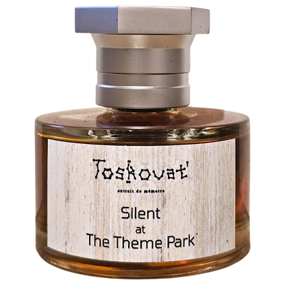 A bottle of Toskovat' 'Silent at The Theme Park' extrait de memoires perfume, featuring amber liquid in a clear glass bottle with a silver-toned metal cap and a light wood-grain textured label.
