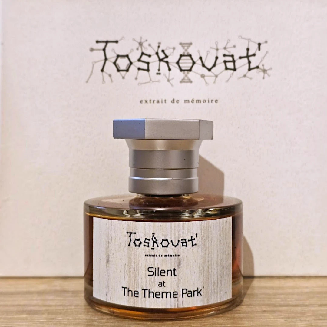 A bottle of Toskóvat 'Silent at The Theme Park' extrait de mémoire perfume or fragrance, featuring a clear glass bottle with amber liquid, a silver hexagonal cap, and a light wood-grain label, set against a matching beige background with the brand name.