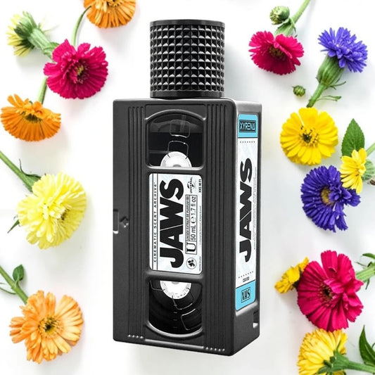 A novelty VHS-shaped bottle or container labeled 'JAWS' in large white letters, surrounded by colorful flowers on a white background. Xyrena jaws | Fragrance Samples UK 1ml 2ml and 5ml perfume samples available 